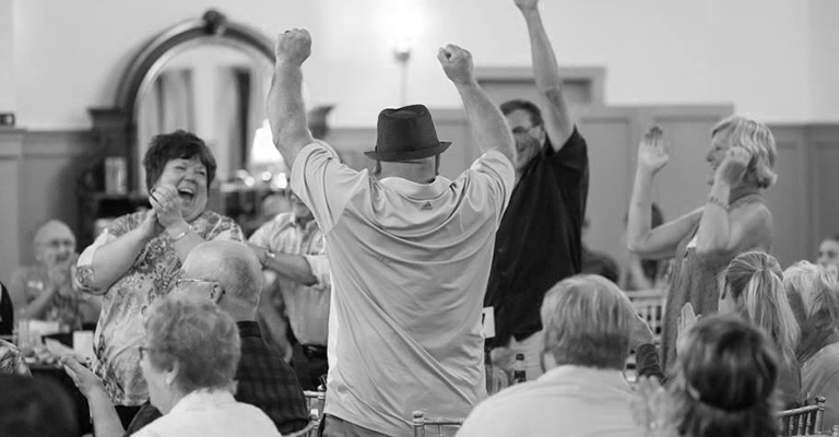 A black and white photo of people sitting in a dining room. A person is standing with their back to the camera and has their arms up in celebration and several others are also standing and clapping